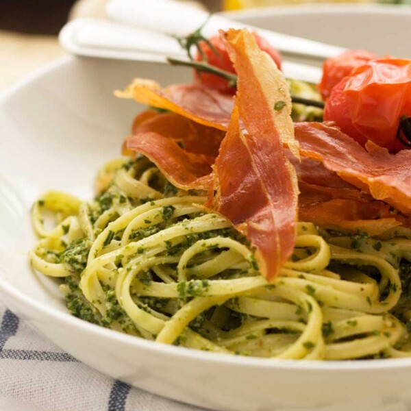 A closeup of a white bowl of kale pesto linguine with cutlery on a blue and white checked tea towel.