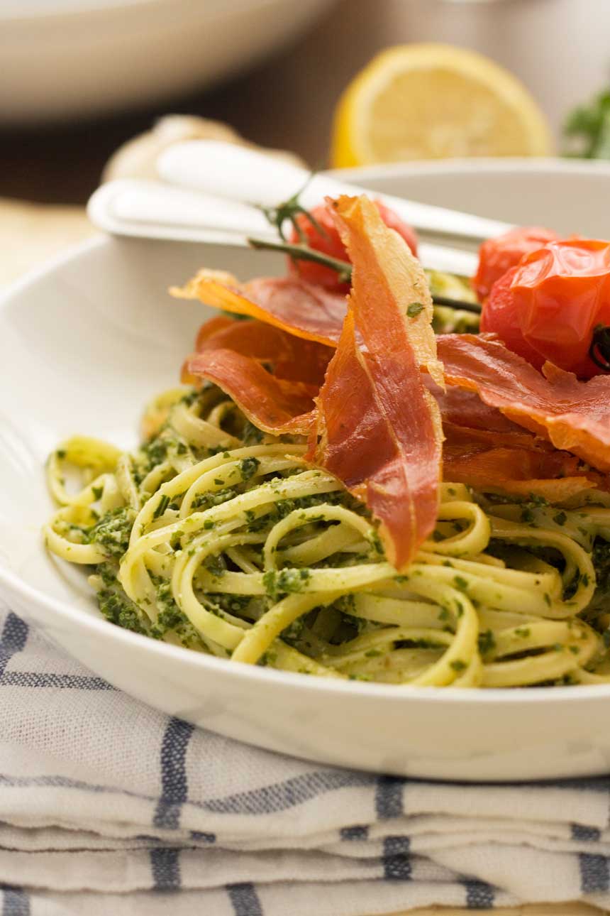 A closeup of a white bowl of kale pesto linguine with cutlery on a blue and white checked tea towel.