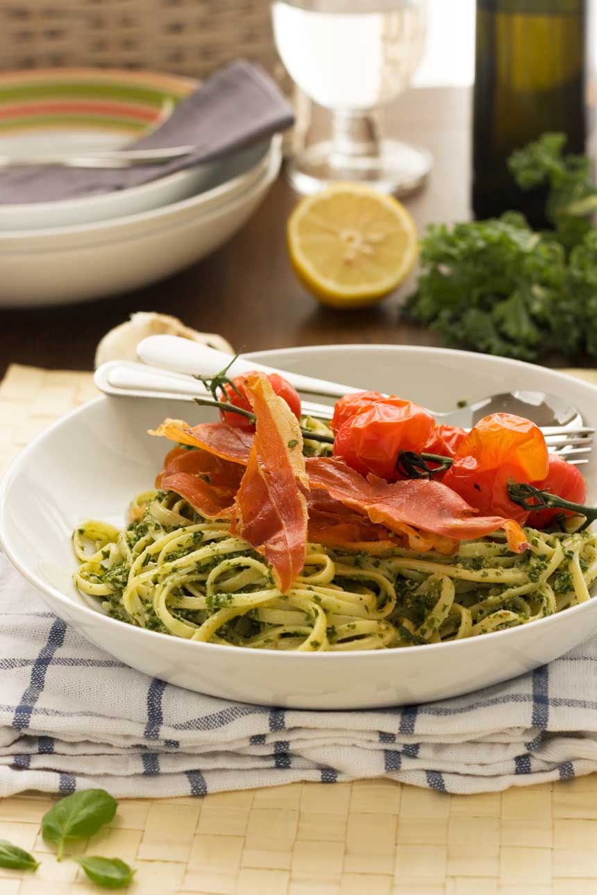 A white bowl of kale pesto linguine with cutlery on a blue and white checked tea towel with a lemon and plates in the background.