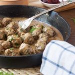 A cast iron pan of Swedish meatballs with a spoon in it and a blue and white checked tea towel