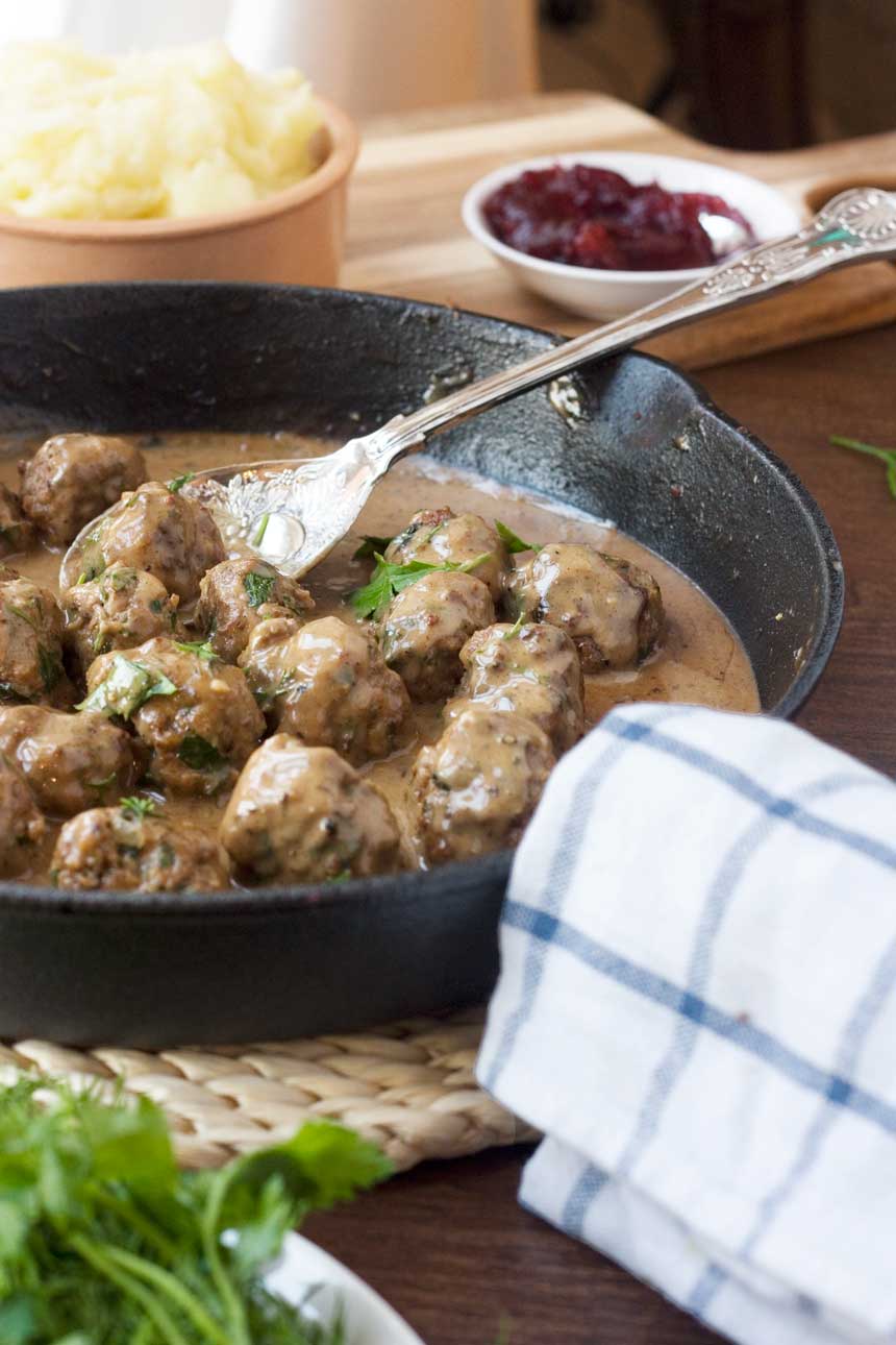 A cast iron pan of Swedish meatballs with a spoon in it and a blue and white checked tea towel