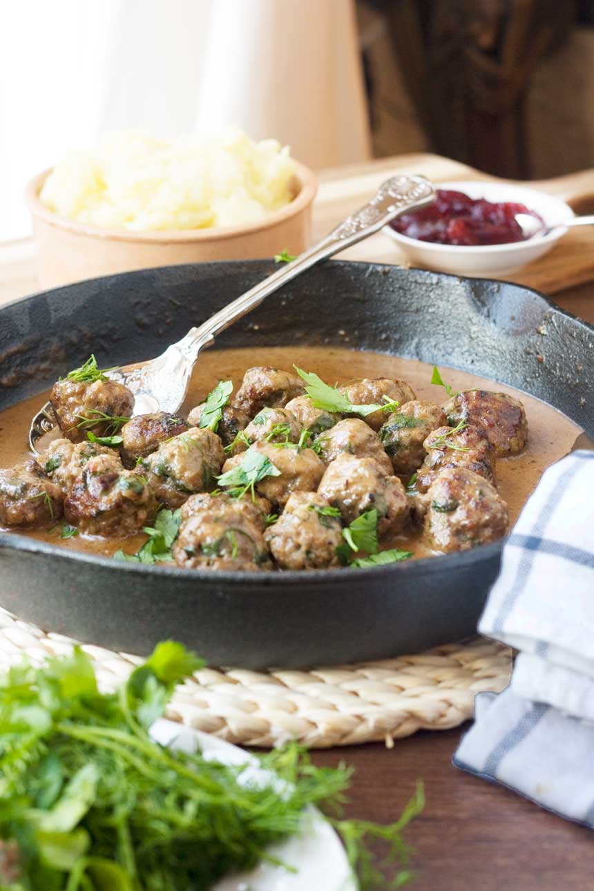A cast iron pan of Swedish meatballs with mashed potatoes and red sauce in the background