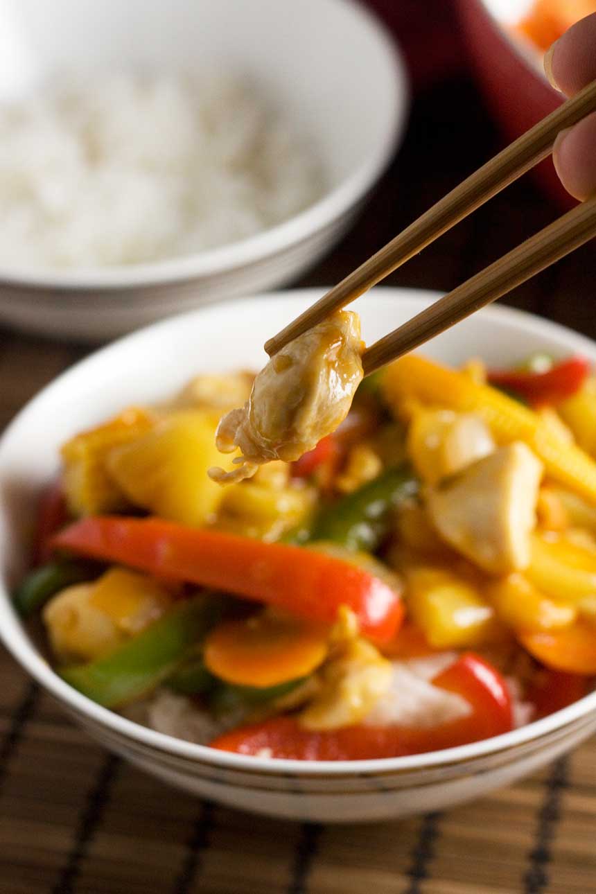 Someone holding up a piece of chicken with chopsticks over a bowl of sweet and sour chicken