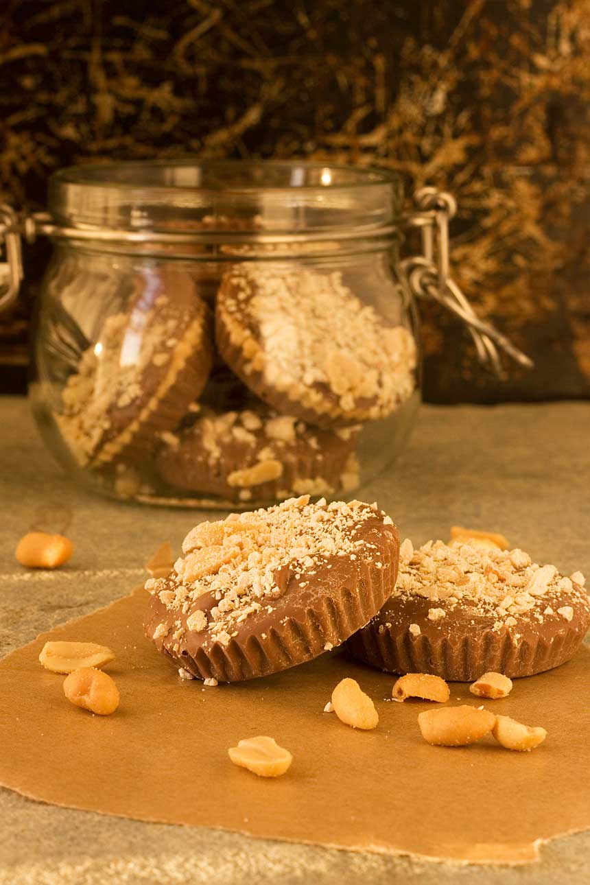 Homemade peanut butter cups on a table with nuts scattered around and with a jar of them behind.