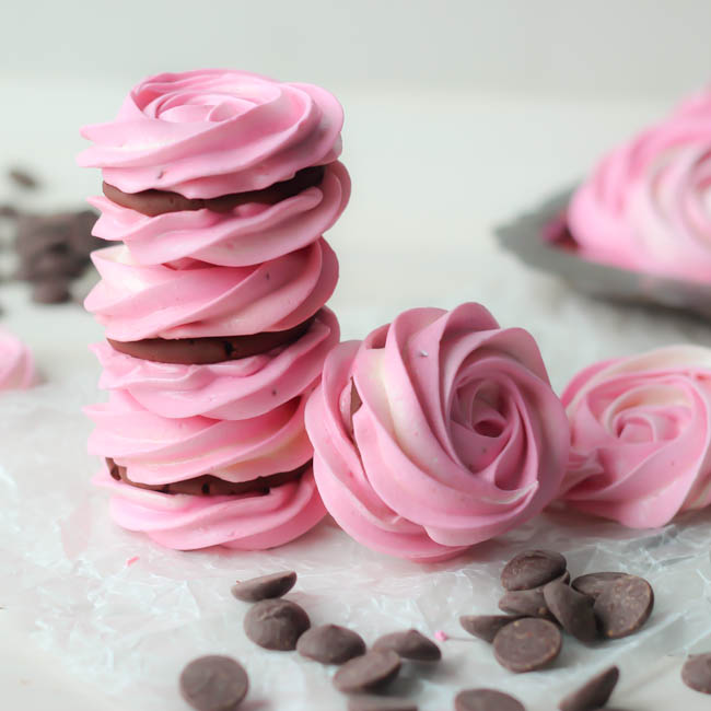 Raspberry meringue sandwiches with whipped dark chocolate ganache piled on baking paper with chocolate chips scattered around.