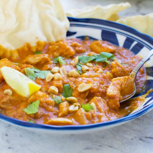 A Thai chicken and butternut squash curry in a blue bowl with peanuts, coriander, poppadoms and a lime wedge, with a spoon