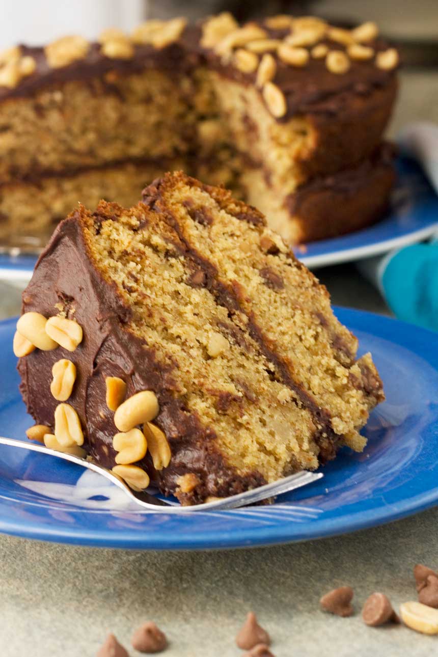 A big slice of peanut butter, banana and chocolate chip cake on a blue plate with a fork with the whole cake in the background.