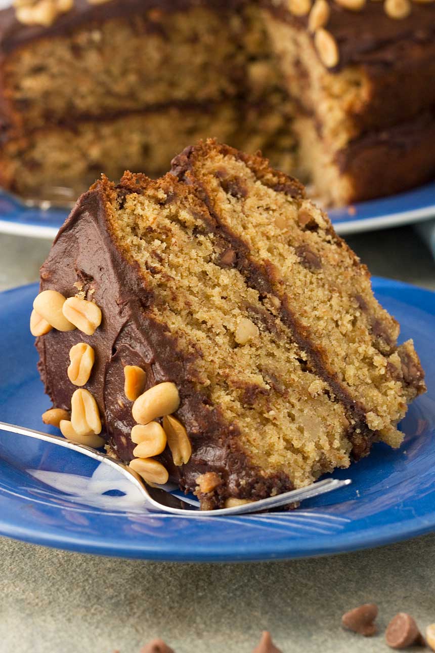 A slice of peanut butter banana choc chip cake on a blue plate with a fork and the rest of the cake in the background.