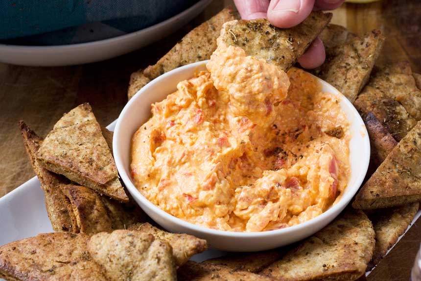 Someone scooping roasted red pepper dip out of a white bowl with Greek pitta chips.