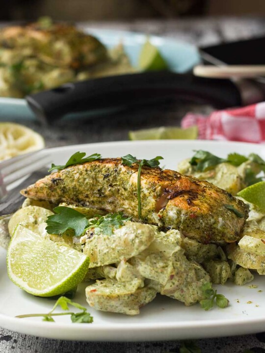 Jerk salmon with yogurt potatoes piled onto a white plate with a fork and a lime wedge with equipment and more food in the background.