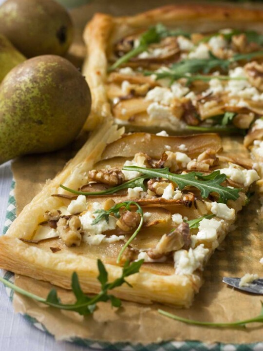 A square of Pear, feta and walnut tart on brown baking paper with more tart in the background and a pear.