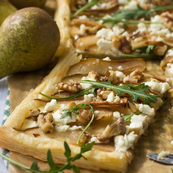 A square of Pear, feta and walnut tart on brown baking paper with more tart in the background and a pear.