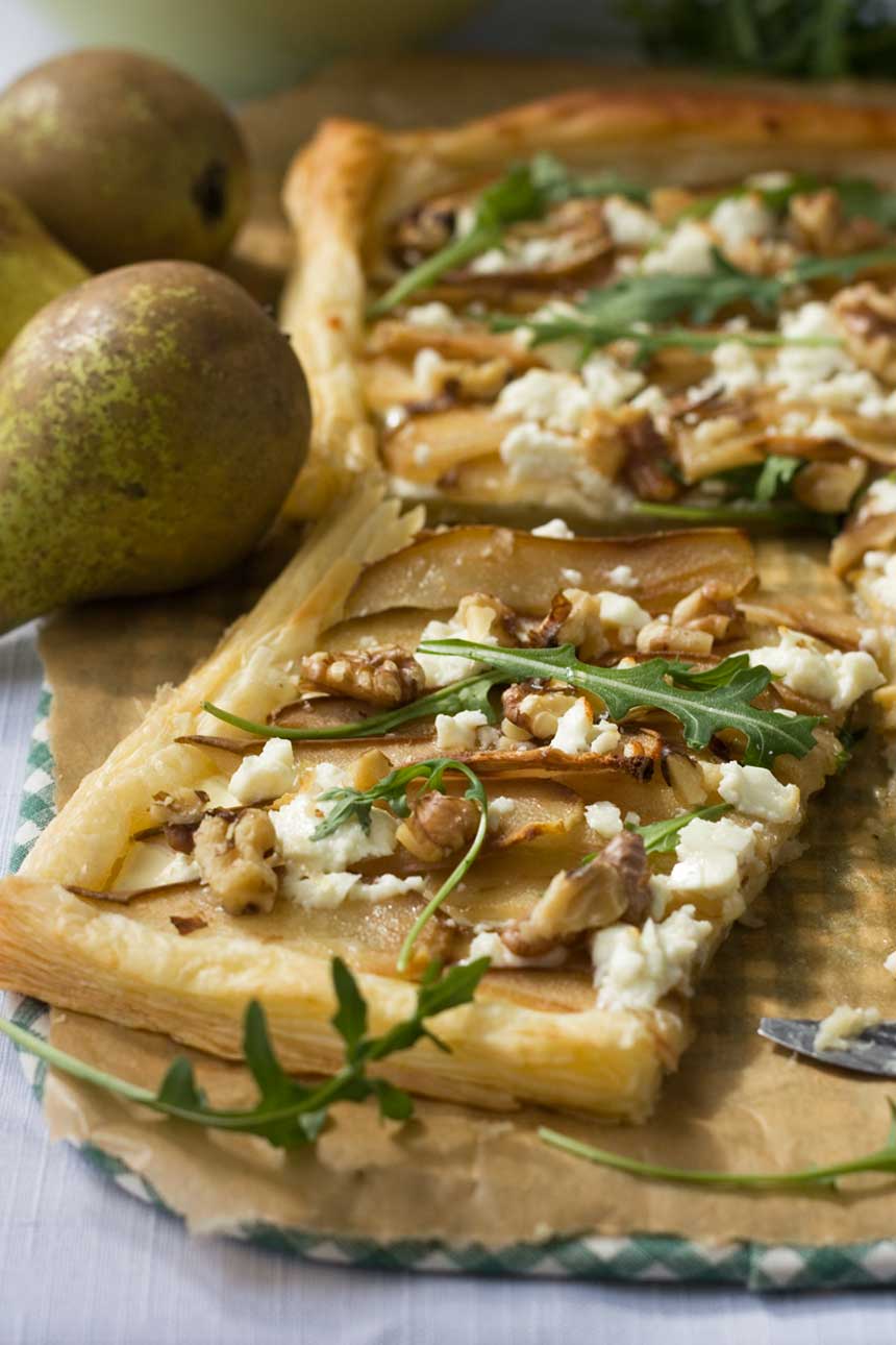 A square of Pear, feta and walnut tart on brown baking paper with more tart in the background and a pear.