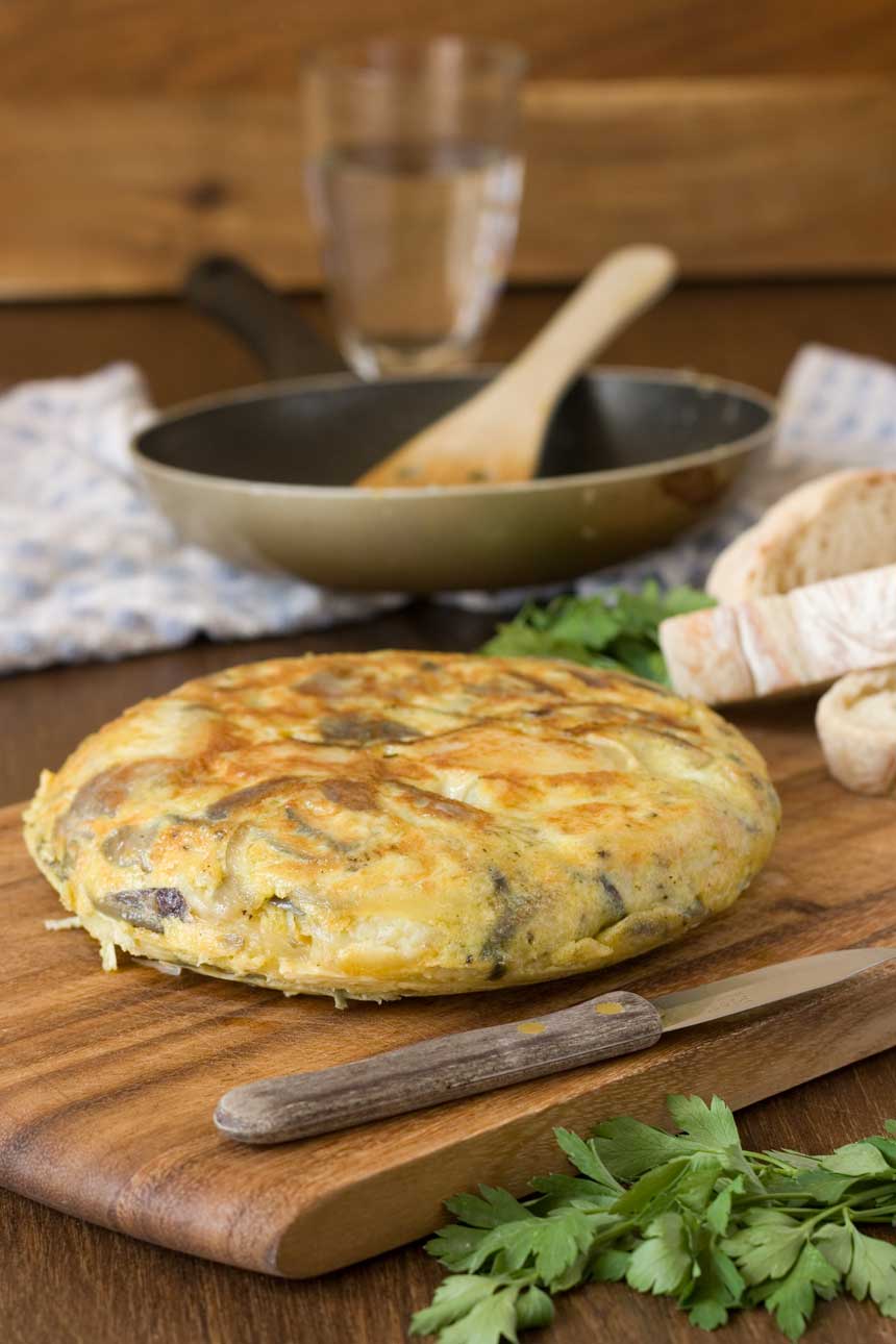 A whole Spanish omelette on a wooden cutting board with a pan in the background