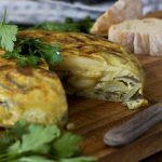 A close up of a Spanish tortilla or omelette with a big slice cut out of it on a cutting board with bread in the background