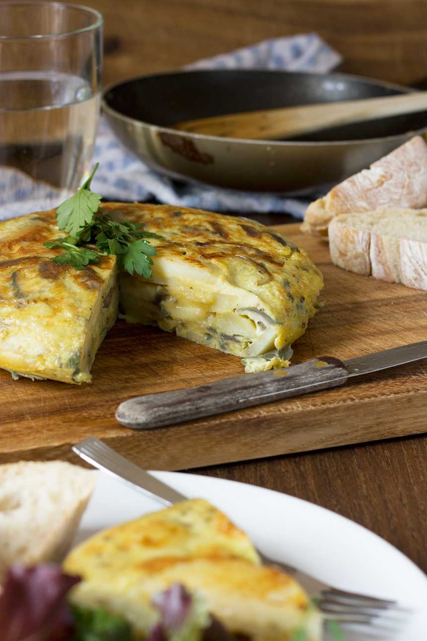 A Spanish omelette on a wooden cutting board with a slice out of it and another piece in the foreground