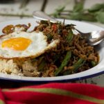 A white plate of Thai basil pork with rice and an egg with a red tea towel in front.