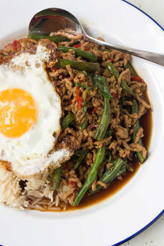 A Thai basil stir-fry from above on a white plate with an egg on top and a spoon on the plate 