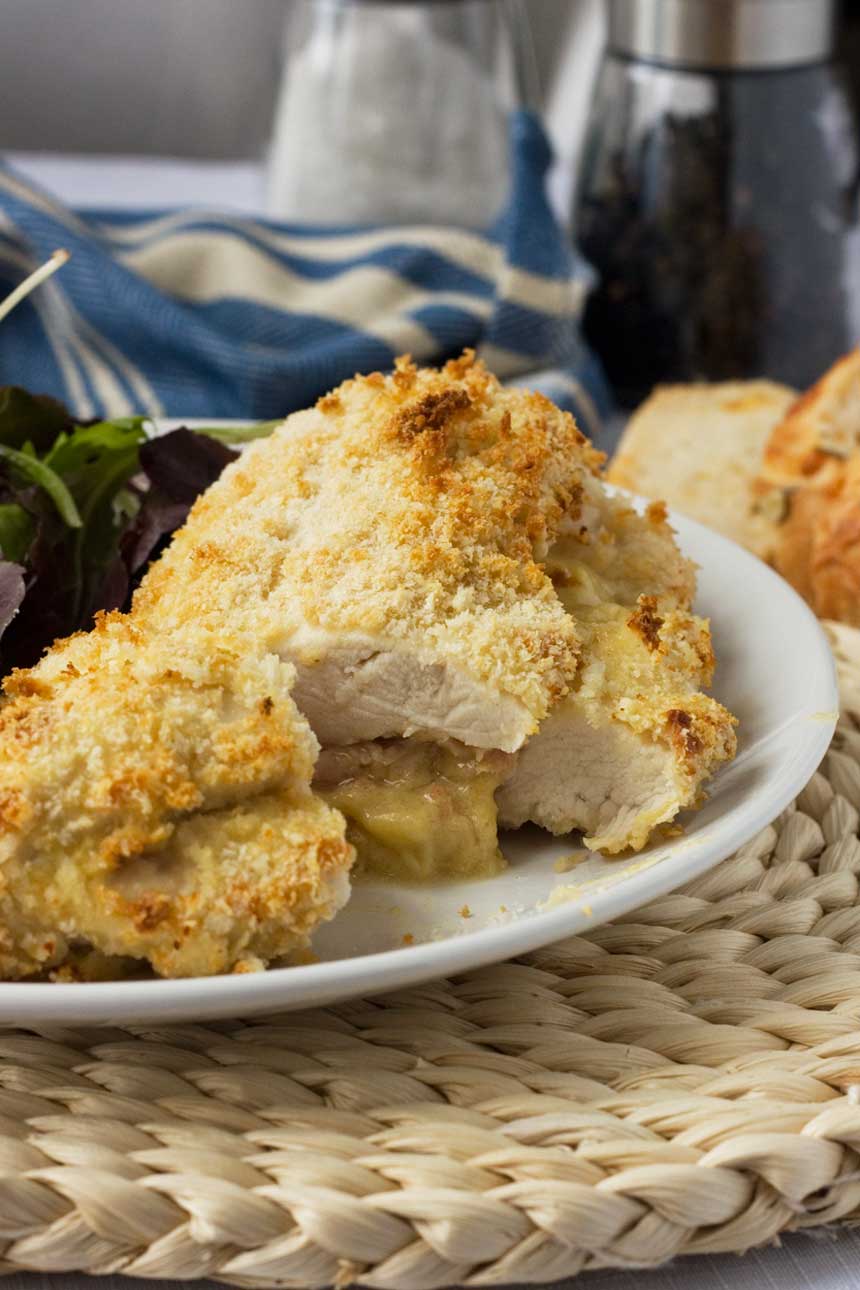 Crispy baked cheese and ham stuffed chicken on a white plate and wicker place mat with a tea towel behind.