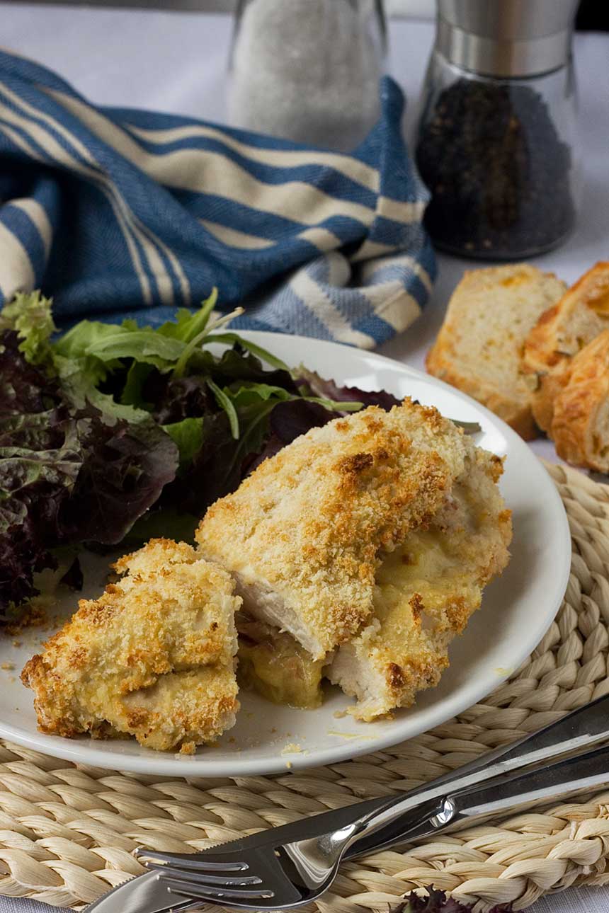 Crispy baked cheese and ham stuffed chicken on a white plate with salad and with a blue tea towel and bread in the background.