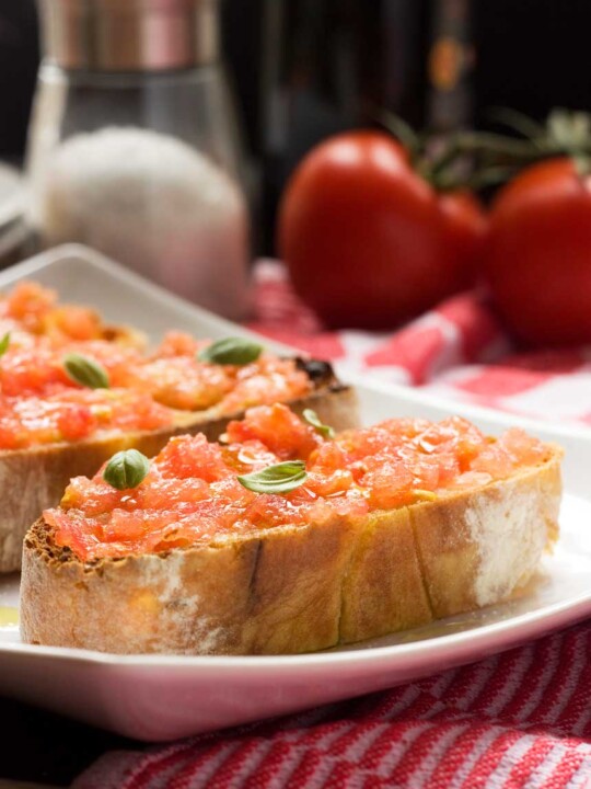 Spanish bruschetta (pan con tomate) close up on a white plate with ingredients behind