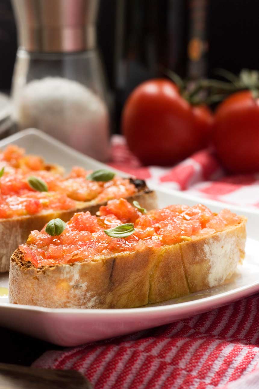 Spanish bruschetta (pan con tomate) close up on a white plate with ingredients behind