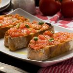 A white rectangular plate with 3 slices of Spanish bruschetta or pan con tomate on it on a red and white tea towel