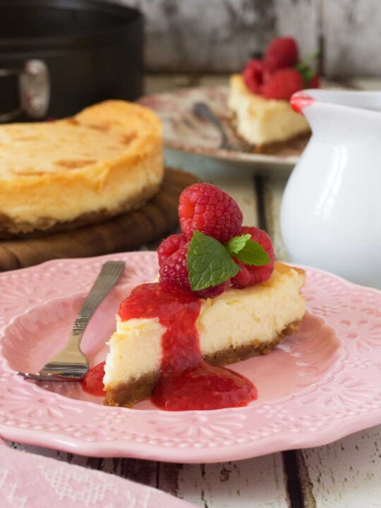 closeup of a piece of White chocolate baked cheesecake with raspberry coulis on a pink plate with the rest of the cheesecake in the background