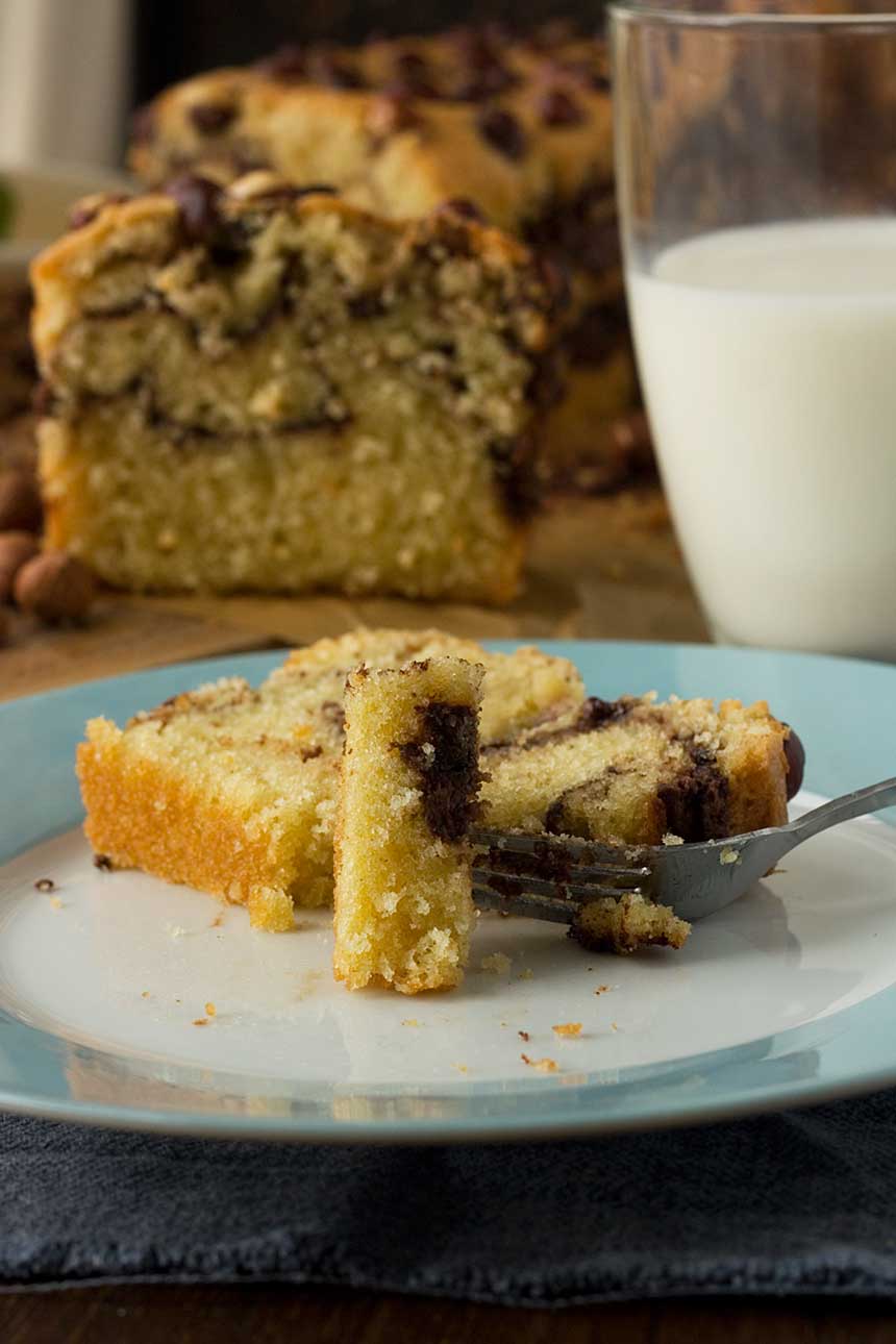 A piece of Nutella swirl loaf cake on a plate with a fork on a white and blue plate and with the rest of the cake and a glass of milk in the background