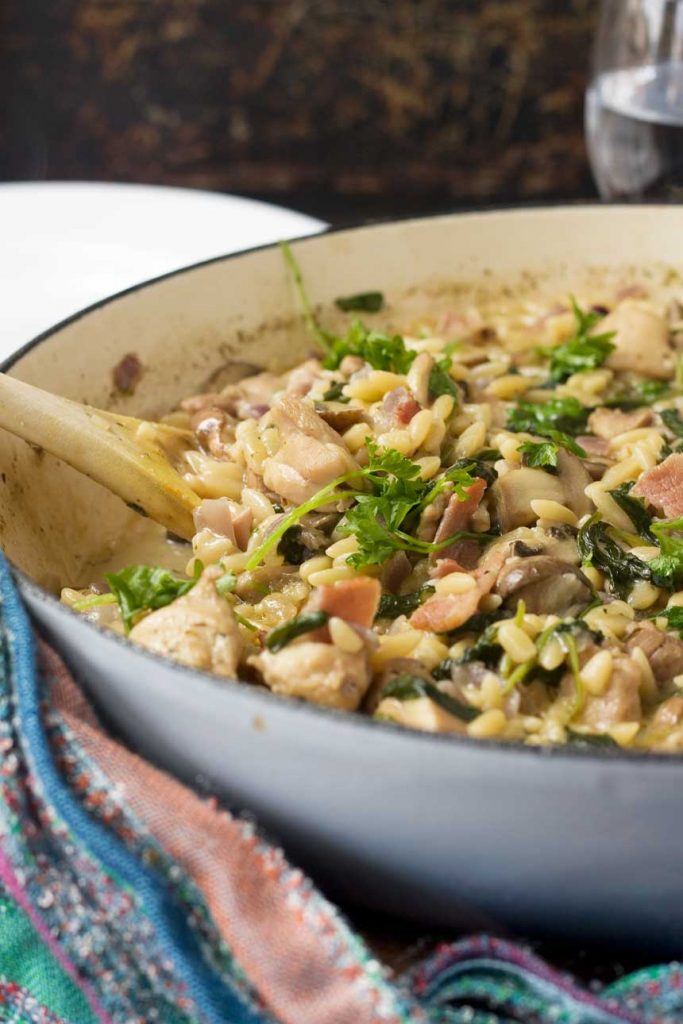Closeup of a one-pot creamy chicken, bacon and mushroom orzo in a blue cast iron pan with a multi-coloured tea towel in front