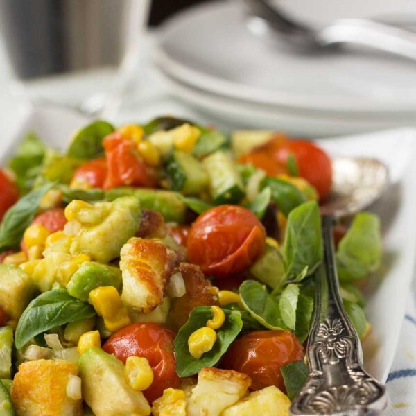 A close up of a corn, avocado, tomato and basil salad with crispy halloumi - there are some white plates in the background