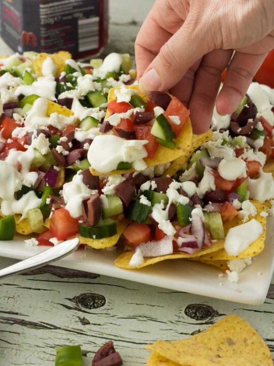 Some holding a loaded Greek nacho above a white plate of them