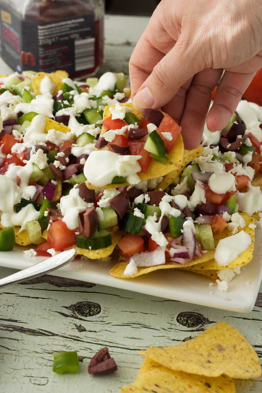 Some holding a loaded Greek nacho above a white plate of them