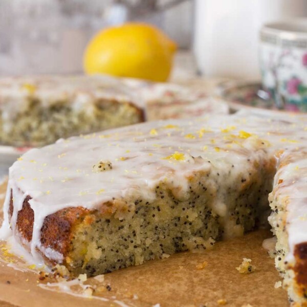 A piece of lemon poppy seed drizzle cake on baking paper with a big piece cut out of it and with ingredients in the background.
