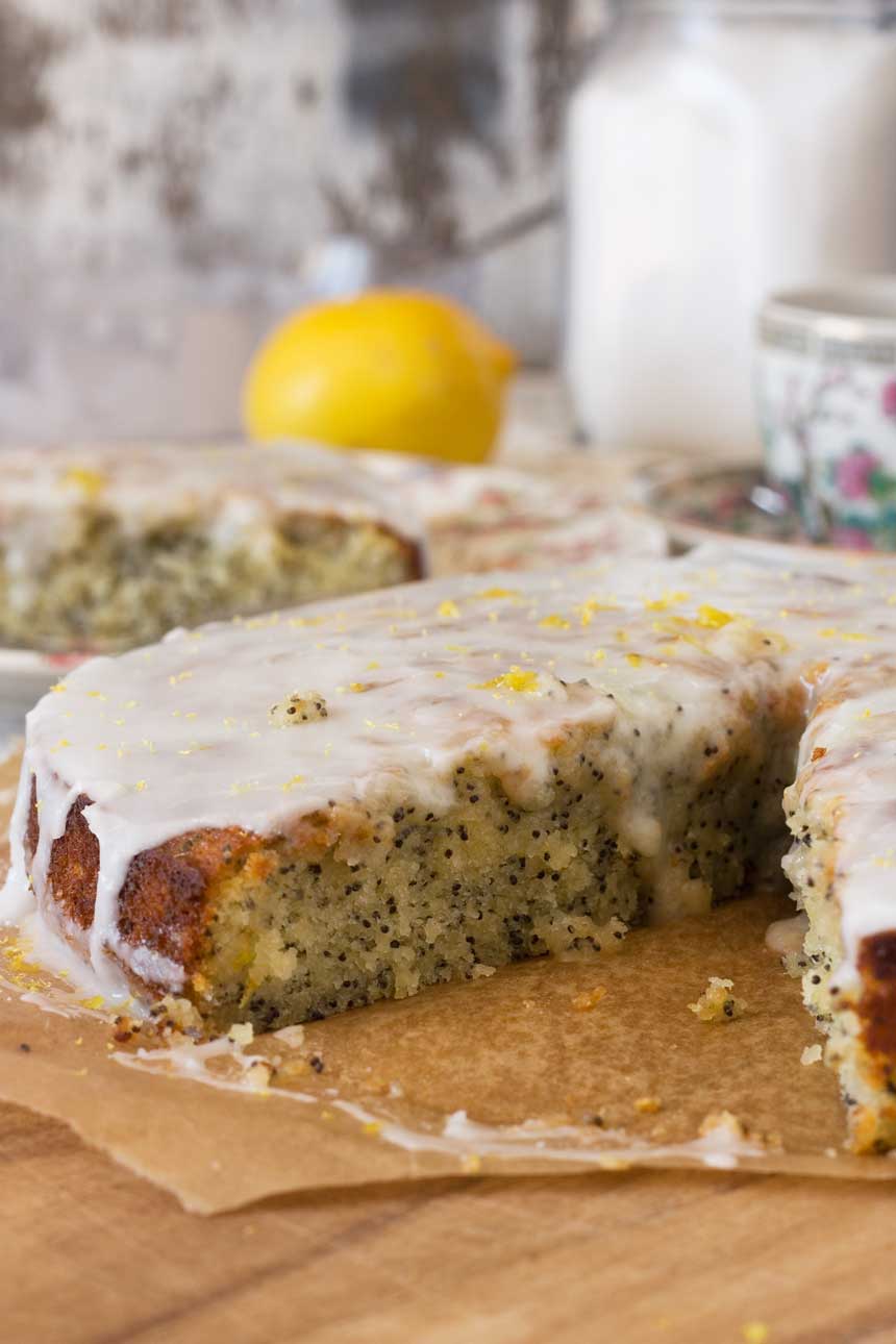 A piece of lemon poppy seed drizzle cake on baking paper with a big piece cut out of it and with ingredients in the background.