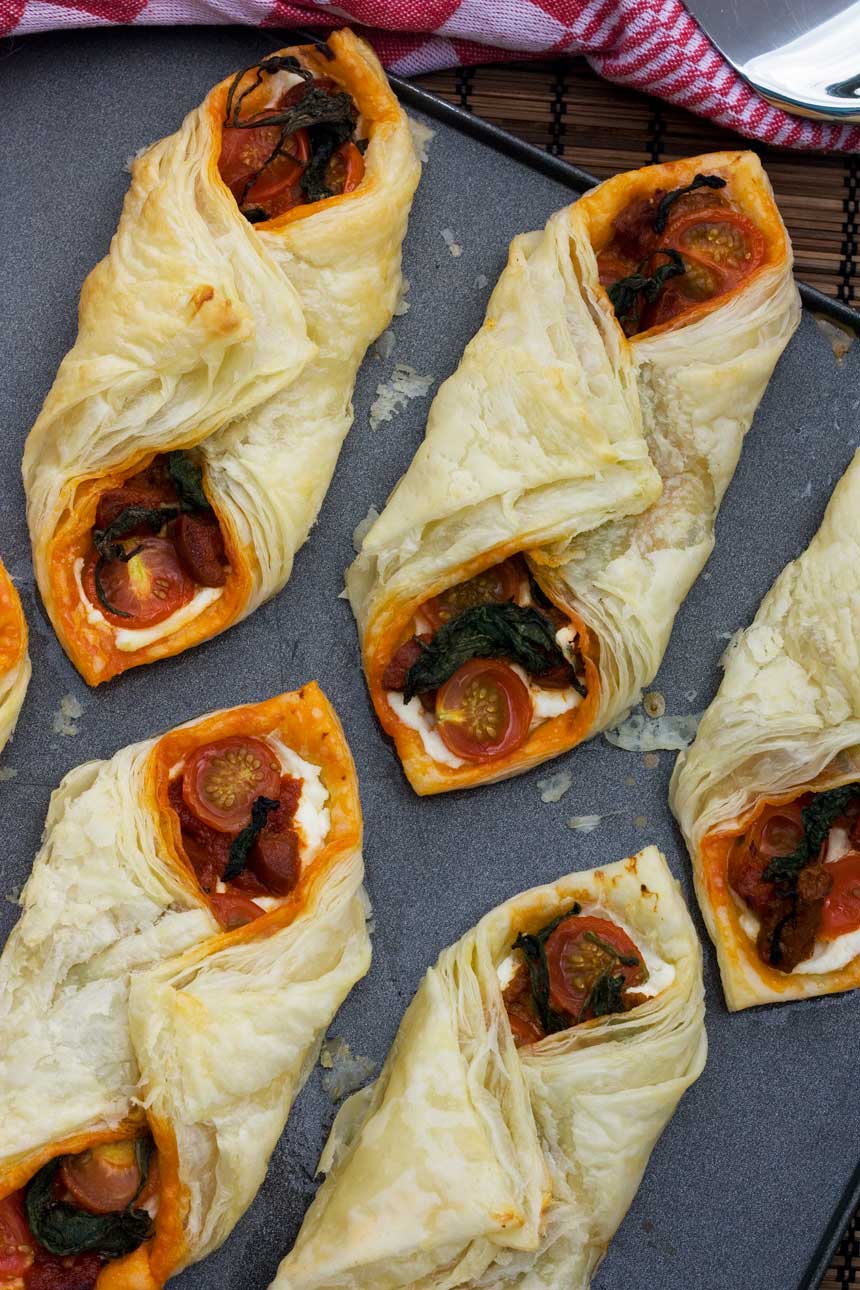 Ricotta, cherry tomato and chorizo puff pastry tarts lined up on a baking tray from above.