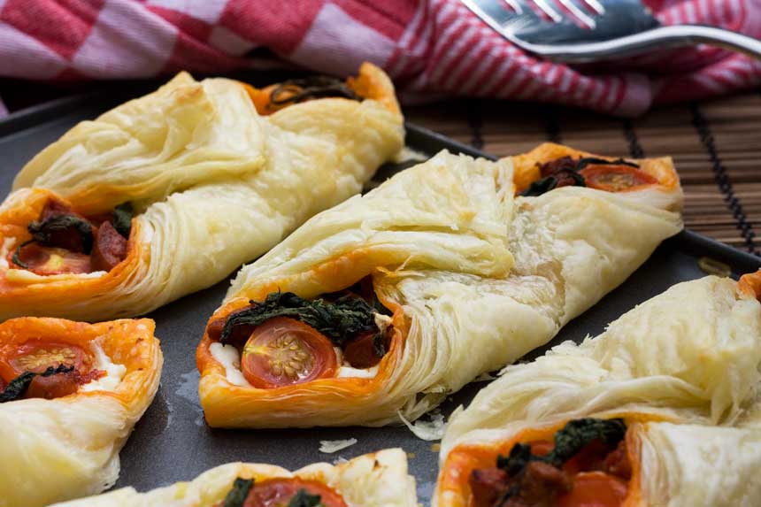 Chorizo and puff pastry tarts in a row on a baking tray with a red tea towel behind.