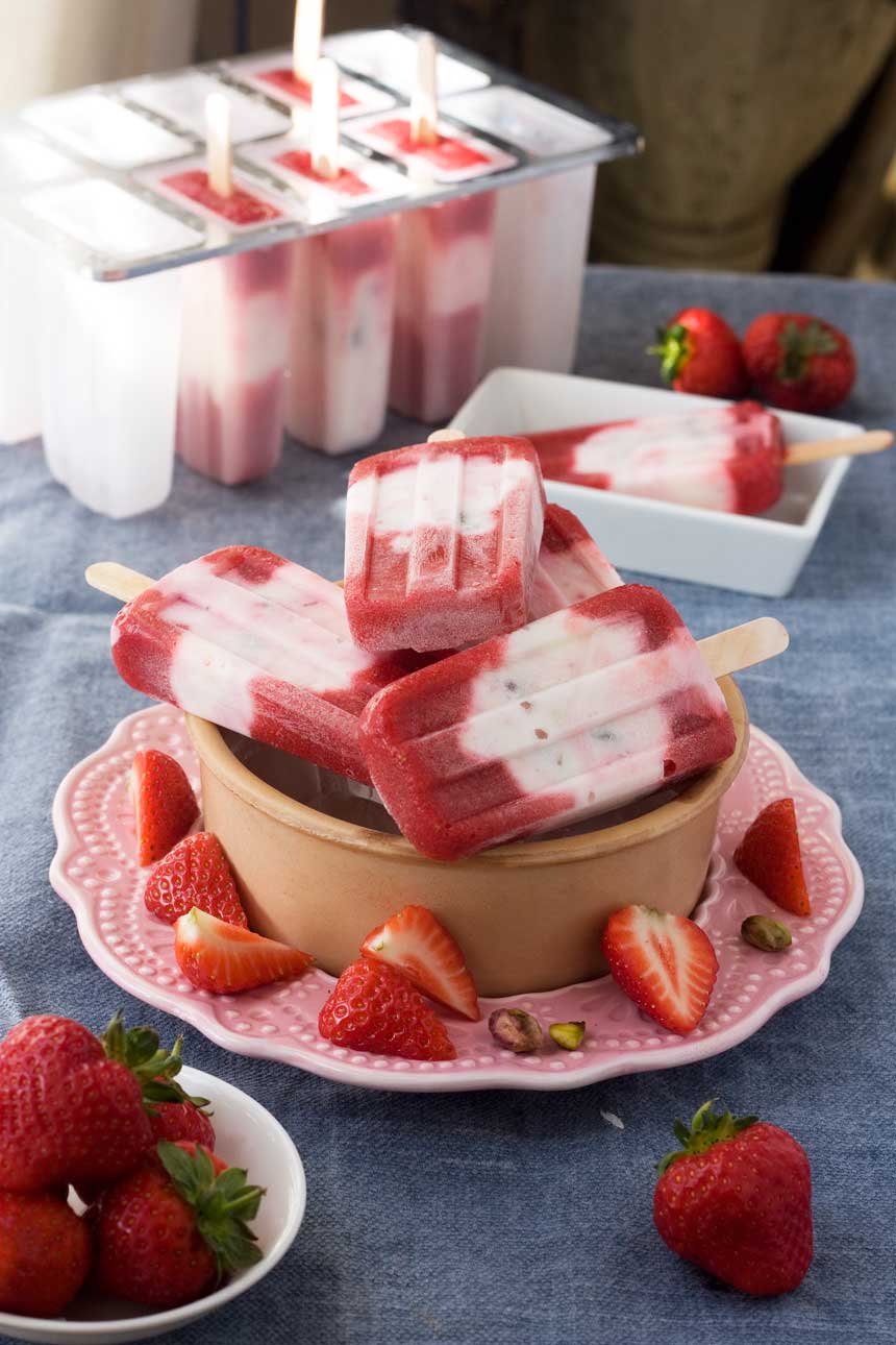 A bowl of strawberry pistachio yoghurt popsicles on a pink plate and blue background with strawberries around it.