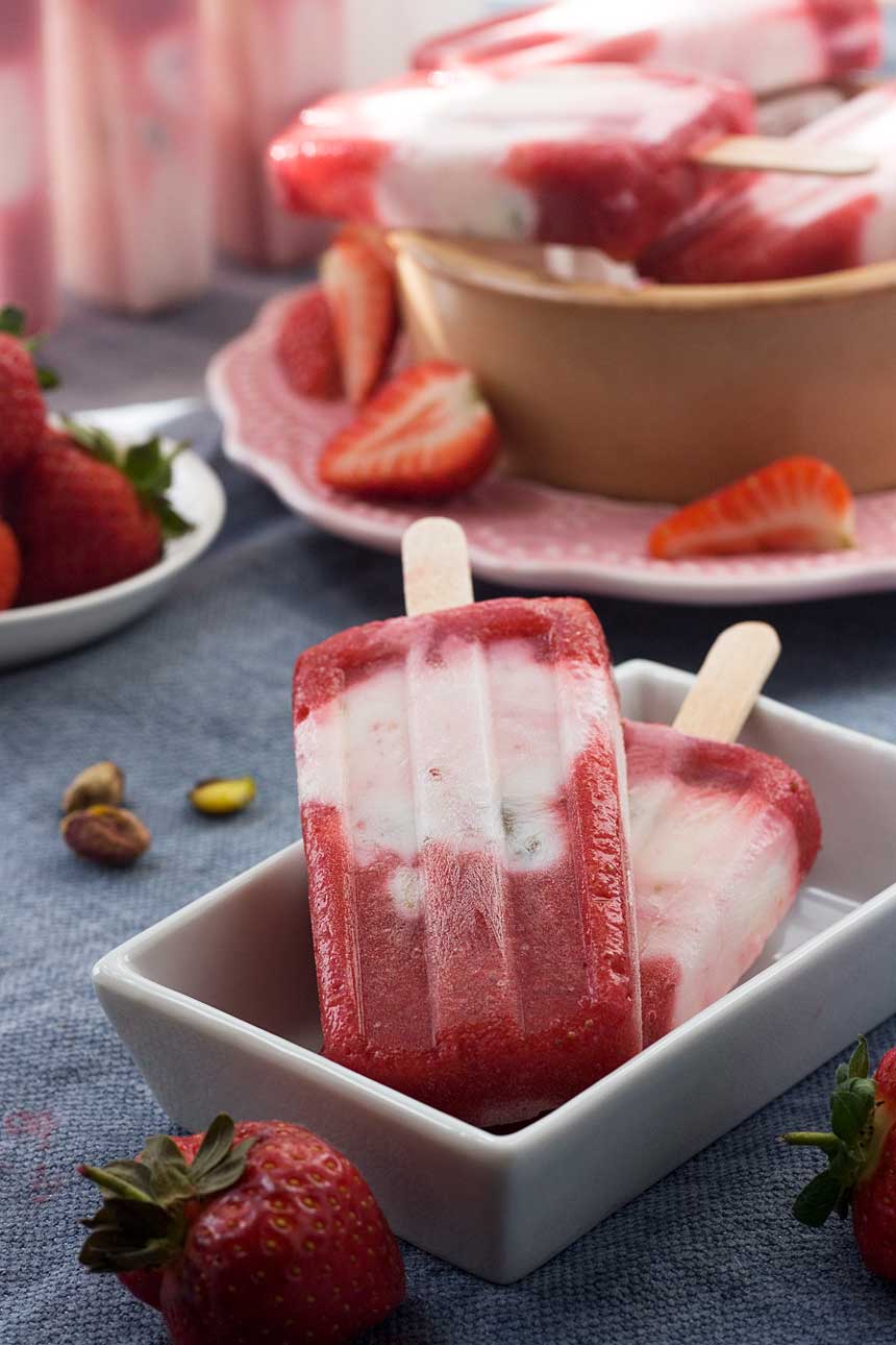 A closeup of strawberry pistachio yoghurt popsicles in a white dish on a blue background and with strawberries in the background.