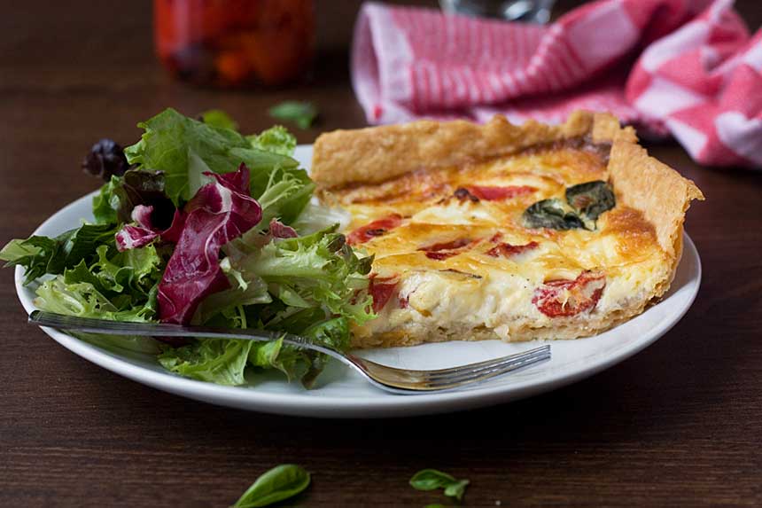 A square slice of feta and red pepper quiche on a white plate with green salad and a fork.