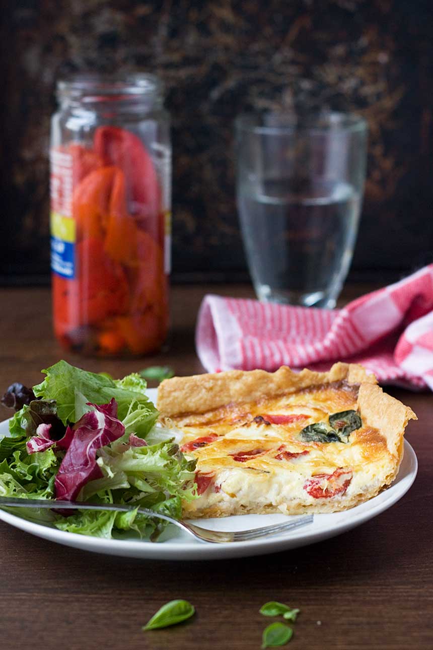 A square slice of easy feta and red pepper quiche on a white plate with green salad and a fork and with ingredients in the background.