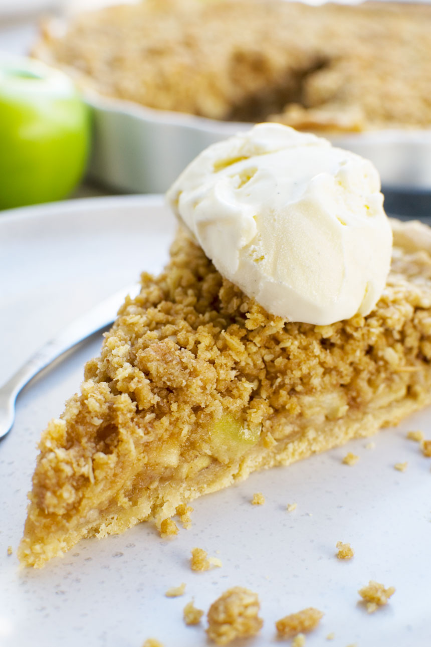 A close up of a piece of easy apple crumble pie on a white plate with ice cream on top and apples in the background