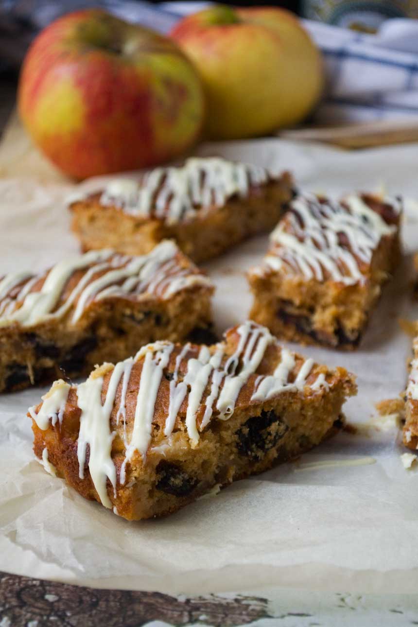 A close up of sticky apple ginger cake bars on baking paper with apples and other ingredients in the background