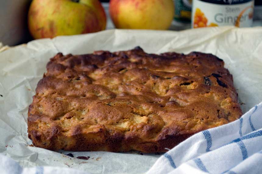 Whole sticky apple ginger cake bars on baking paper with apples and other ingredients in the background