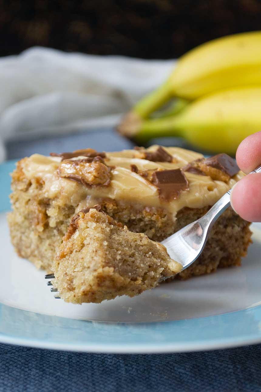 Someone eating A piece of banana cake with peanut butter frosting with a fork on a white plate with bananas in the background.