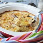 A red oval baking dish of creamy garlic parmesan potatoes with a spoon in it and a multi-coloured stripy tea towel in the foreground