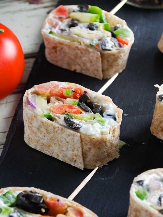 Closeup of Greek tortilla pinwheels on a black slate platter and with a big tomato on the left.