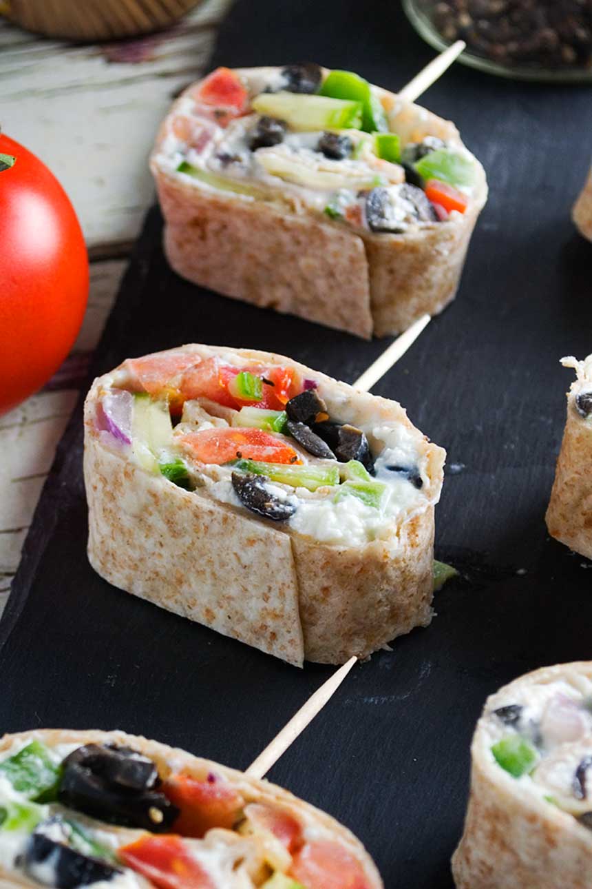 Closeup of Greek tortilla pinwheels on a black slate platter and with a big tomato on the left.