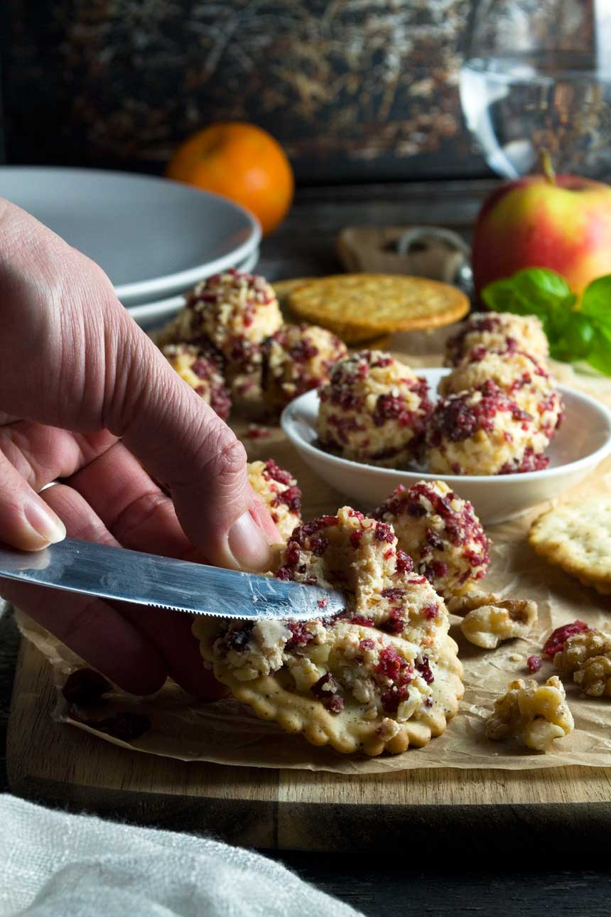 Someone spreading cranberry and walnut cheese truffles onto crackers, with more crackers and truffles in the background.