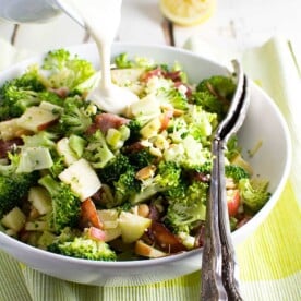 Someone pouring dressing over a bowl of broccoli, apple and bacon salad on a green cloth and with serving spoons in it.
